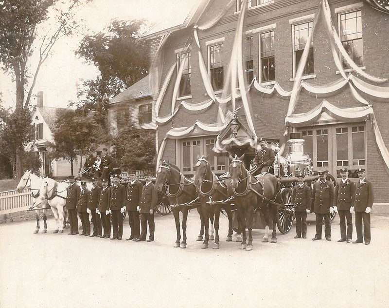 Historical image of a 2 story brick building decorated with ribbons with officers standing and on horseback posing in front.